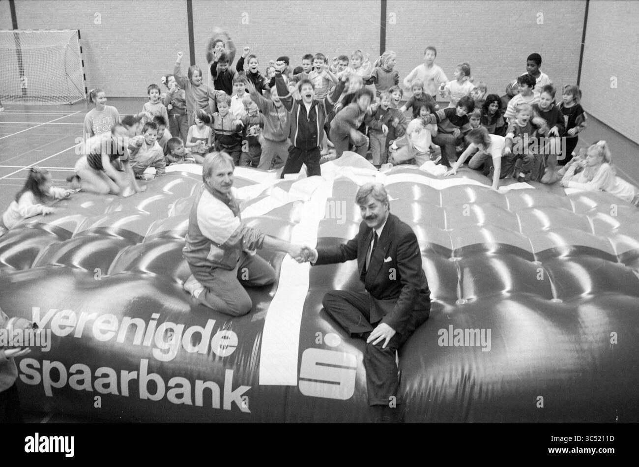 Consegnare il castello rimbalzante VSB a Speliade Spaarnehal, Nuts, United Savings Banks, Giochi e giocattoli, Haarlem, FIE Carelsenplein, Paesi Bassi, 03-01-1989 Whizgle News, Dutch Desk, Paesi Bassi, 1950-2000 Un gruppo di bambini eccitati celebra intorno a due uomini che stringono la mano su una grande struttura gonfiabile, simboleggiando una partnership o un evento, con espressioni gioiose e movimenti energici che riempiono lo spazio. Foto Stock