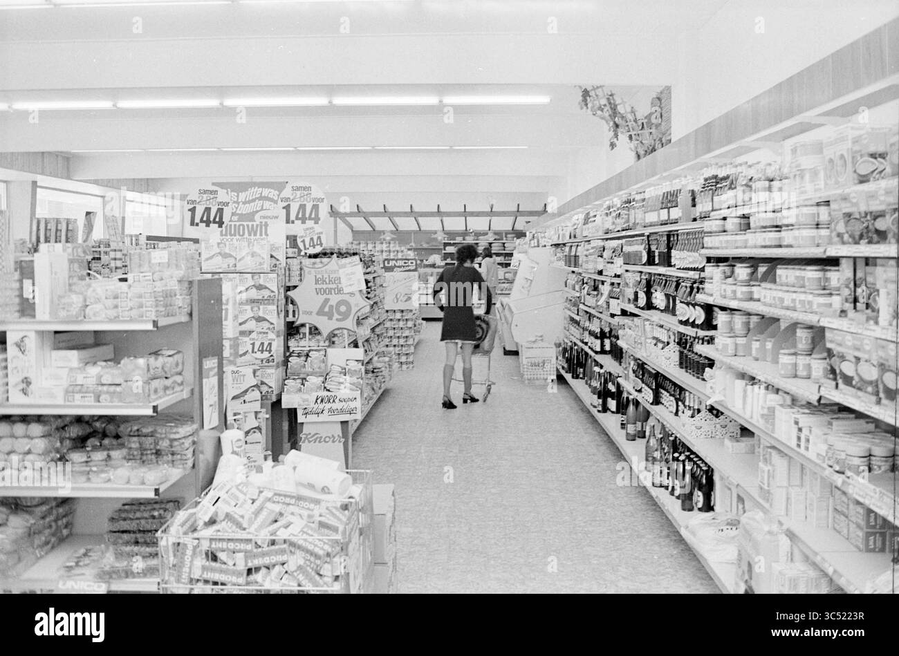 Int Supermercato, interni, 30-08-1971 Whizgle News, Dutch Desk, Paesi Bassi, 1950-2000 un corridoio in un negozio di alimentari presenta scaffali ordinatamente disposti pieni di vari prodotti confezionati, mentre un acquirente naviga tra le esposizioni, apparentemente focalizzato sulle loro selezioni in mezzo alla segnaletica promozionale. Foto Stock