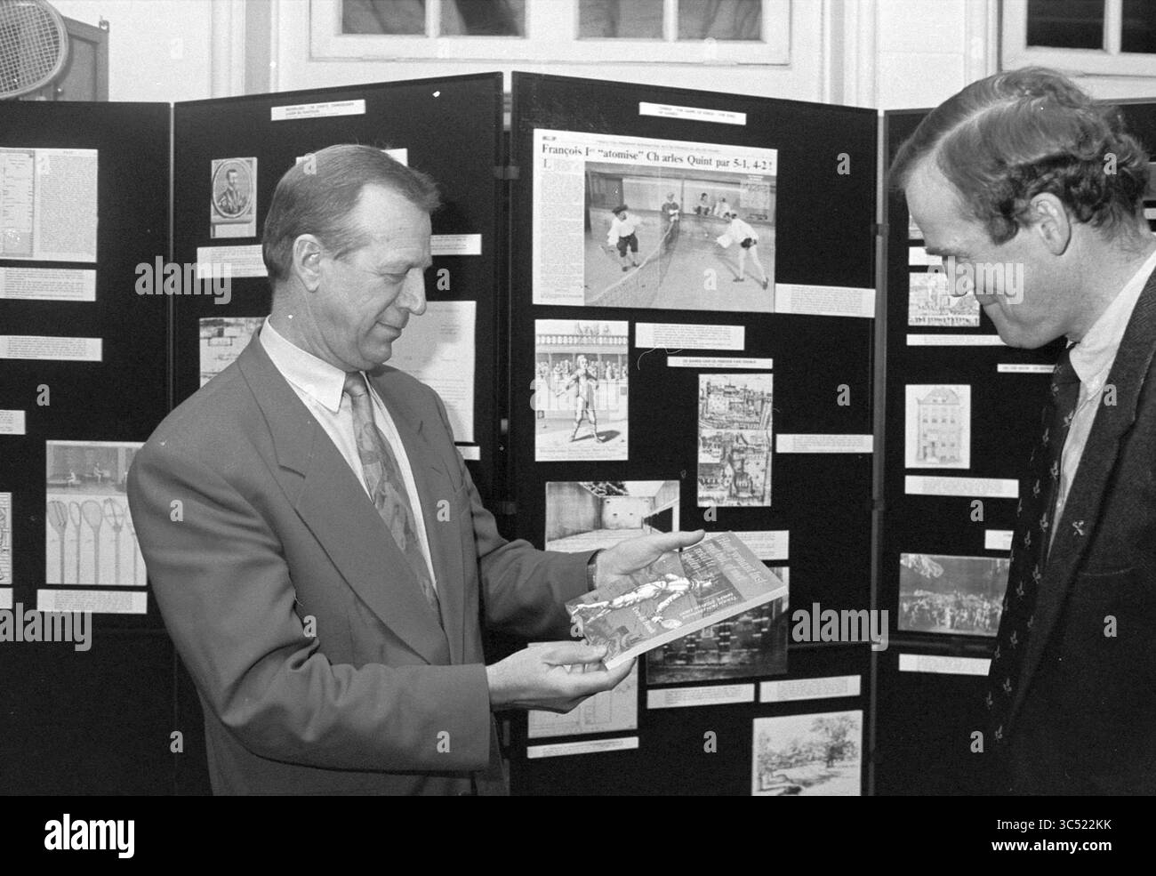 Presentazione del libro a Roosenboom, 18-11-1993 Whizgle News, Dutch Desk, Paesi Bassi, 1950-2000 due uomini si impegnano in conversazione, uno tiene un libro mentre lo mostra all'altro, che sembra essere interessato. Dietro di loro, una collezione di bacheche mostra vari articoli e immagini relativi allo sport, evidenziando momenti e traguardi storici. Foto Stock