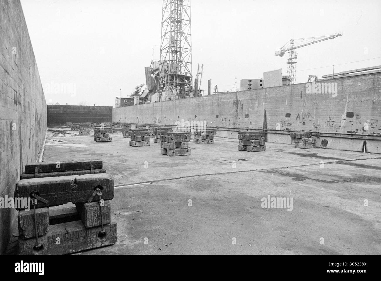 Cantiere Stapel Spaarndam, Spaarndam, Ships, 15-11-1978 Whizgle News, Dutch Desk, i Paesi Bassi, 1950-2000 una scena industriale caratterizzata da un'ampia area portuale vuota circondata da pareti di cemento. Diversi blocchi pesanti sono disposti ordinatamente a terra, con gru e una grande struttura visibile sullo sfondo, suggerendo lavori di costruzione o riparazione in corso nelle vicinanze. L'atmosfera è squallida e funzionale, mettendo in risalto le dimensioni dello spazio. Foto Stock