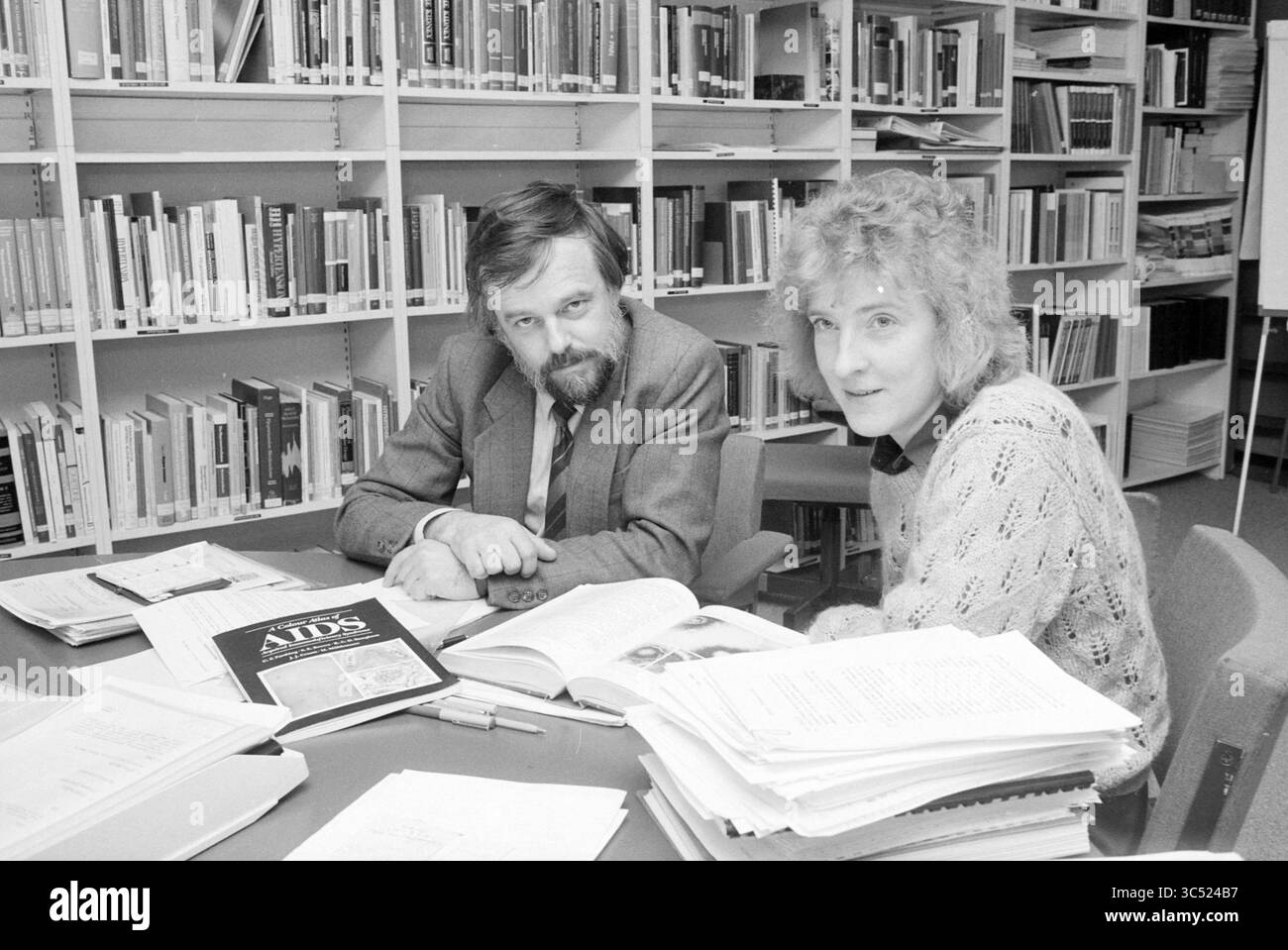 Numero di persone nei vari dipartimenti MSD, MSD, 13-04-1989 Whizgle News, Dutch Desk, Paesi Bassi, 1950-2000 due persone sono sedute ad un tavolo disordinato pieno di libri e documenti relativi alla ricerca sull'AIDS. Sono impegnati in una conversazione, con una persona che guarda direttamente lo spettatore, mentre l'altra sembra esaminare i materiali. Le librerie ricche di numerosi titoli formano lo sfondo, a indicare un ambiente accademico. Foto Stock
