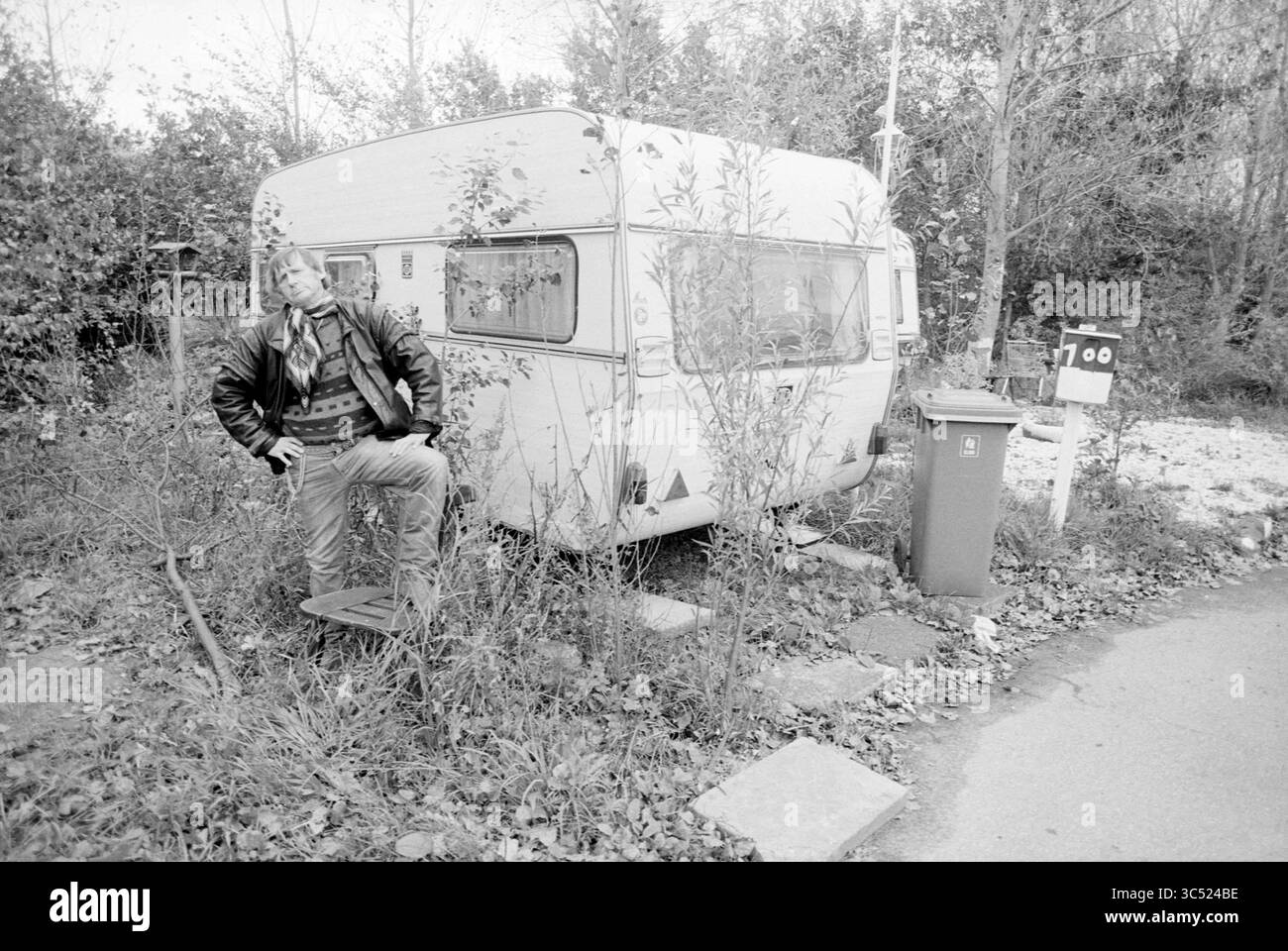 Sjors la Moine, campo carovaniere, IJmuiden, IJmuiden, Paesi Bassi, 06-10-1993 Whizgle News, Dutch Desk, Paesi Bassi, 1950-2000 Un uomo in una giacca di pelle si appoggia casualmente contro una carovana intemprata circondata da vegetazione sovrastata, con una cassetta postale e un cestino nelle vicinanze, suggerendo un ambiente poco chiaro ma rustico. Foto Stock