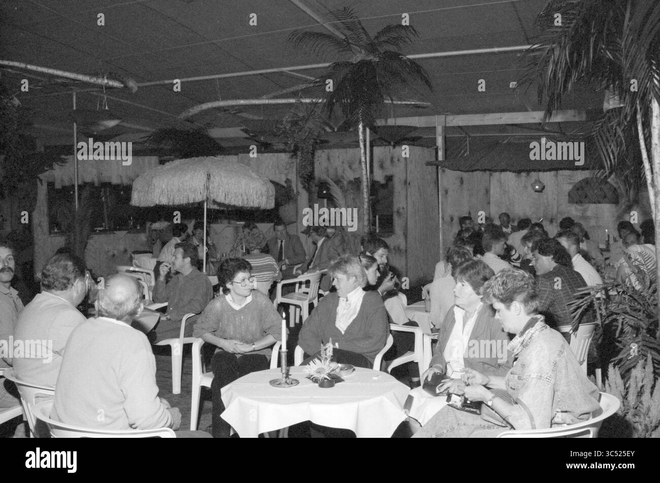 Festa in stile tropicale, 00-08-1987 Whizgle News, Dutch Desk, The Netherlands, 1950-2000 Un vivace raduno si svolge in un luogo a tema tropicale, con gli ospiti seduti ai tavoli circondati da palme e decorazioni in paglia. Le conversazioni fluiscono tra i diversi gruppi, creando un'atmosfera calda e invitante. Foto Stock Festa in stile tropicale, 00-08-1987 Whizgle News, Dutch Desk, The Netherlands, 1950-2000 Un vivace raduno si svolge in un luogo a tema tropicale, con gli ospiti seduti ai tavoli circondati da palme e decorazioni in paglia. Le conversazioni fluiscono tra i diversi gruppi, creando un'atmosfera calda e invitante. Foto Stock