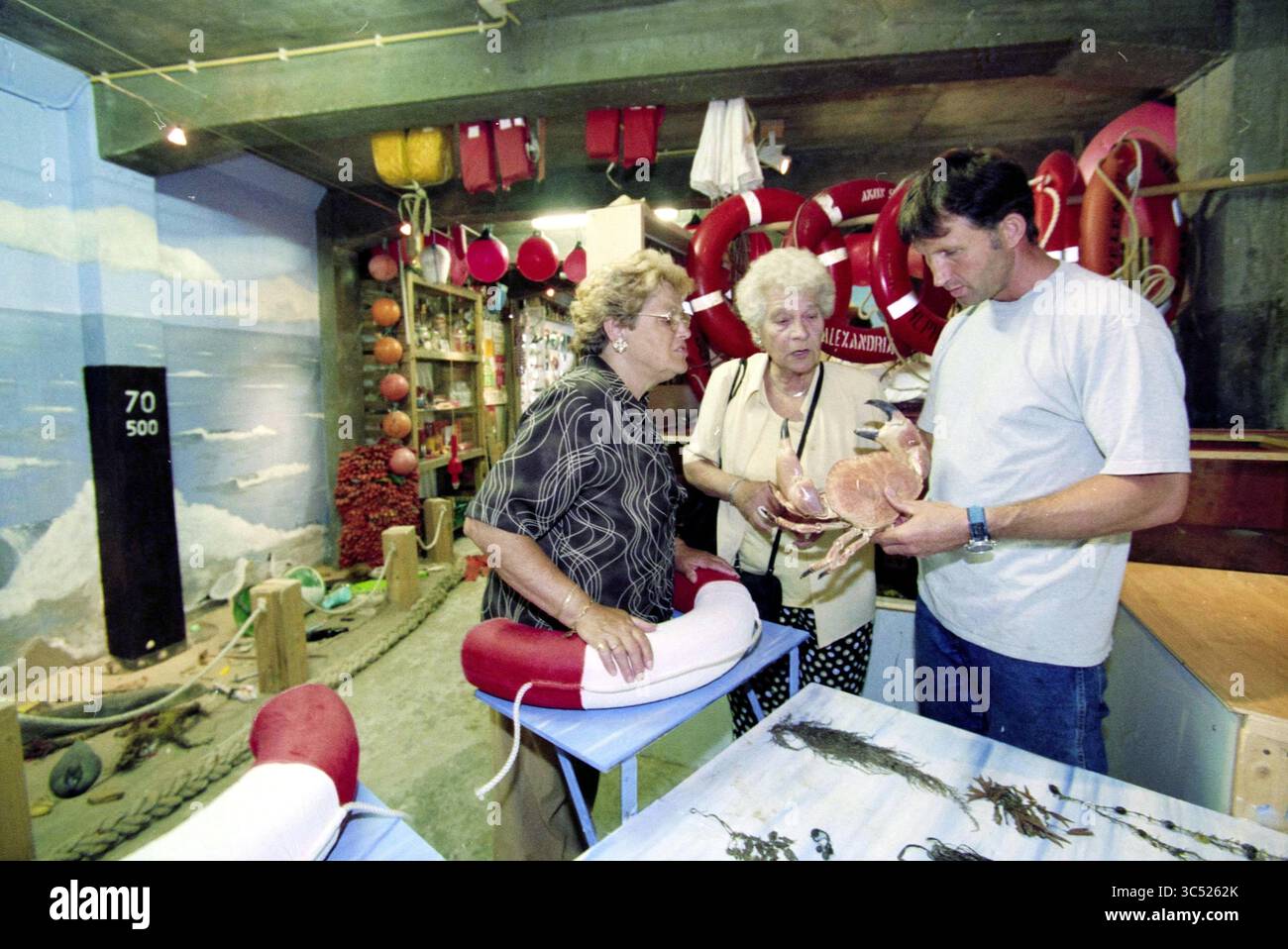 Beachcombers Museum, Zandvoort, Zandvoort, 06-08-2000 Whizgle News, Dutch Desk, i Paesi Bassi, 1950-2000 anni, in un ambiente accogliente e a tema nautico, due donne più anziane si confrontano con un uomo che mostra loro oggetti di interesse. Lo sfondo e' adornato da conservatori di vita, attrezzature sportive e una scena dipinta di onde oceaniche, creando un'atmosfera vibrante ma nostalgica. Foto Stock