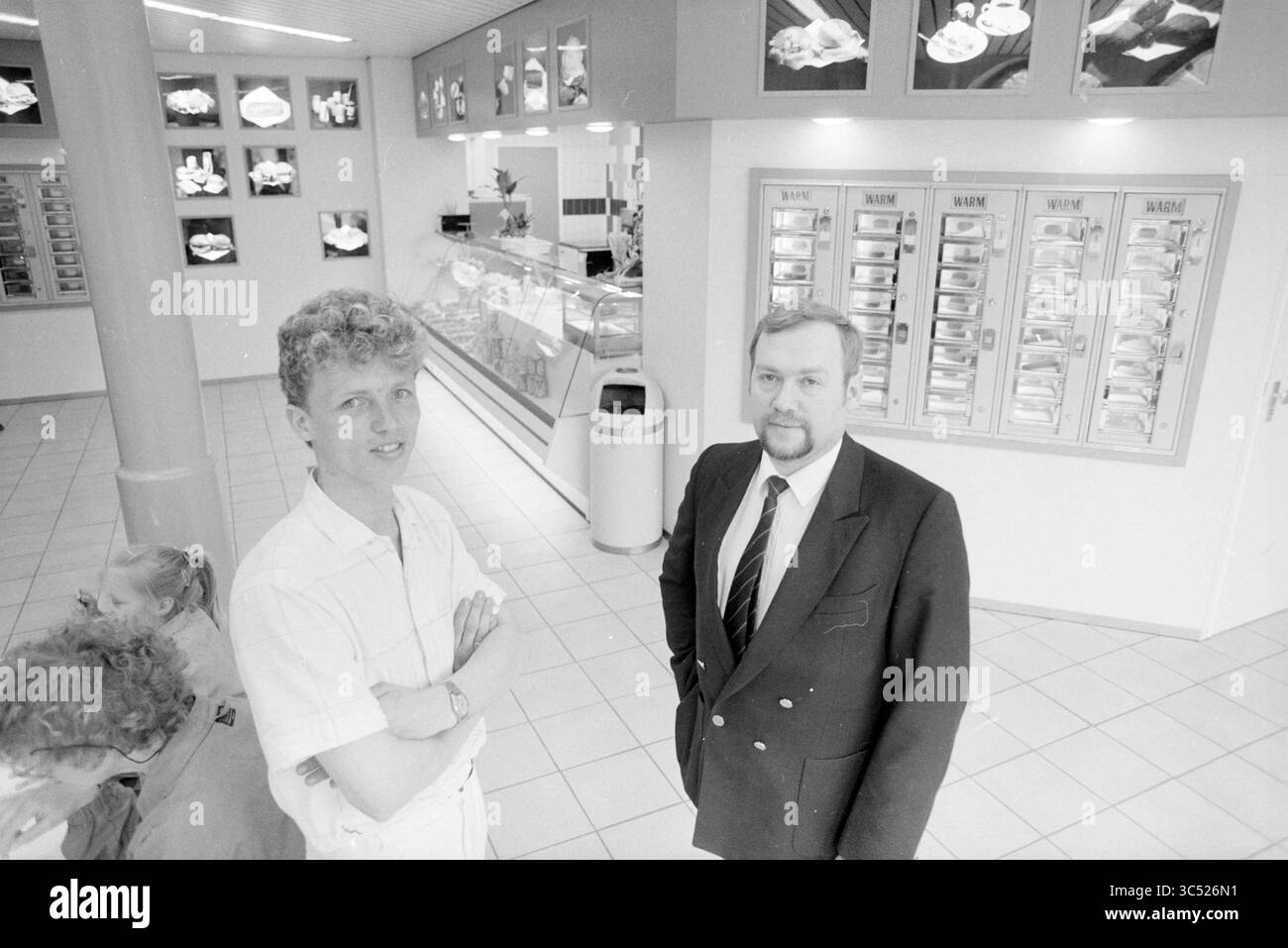 Dipendente e proprietario dello snack bar, 00-05-1987 Whizgle News, Dutch Desk, Paesi Bassi, 1950-2000 due uomini si trovano in un ristorante luminoso e moderno. Uno è vestito in modo informale, mentre l'altro indossa un abito formale. Le pareti sono adornate da menu' che presentano vari piatti e una vetrina piena di cibo e' visibile sullo sfondo. Alcuni clienti possono essere visti nello spazio, creando un'atmosfera vivace. Foto Stock