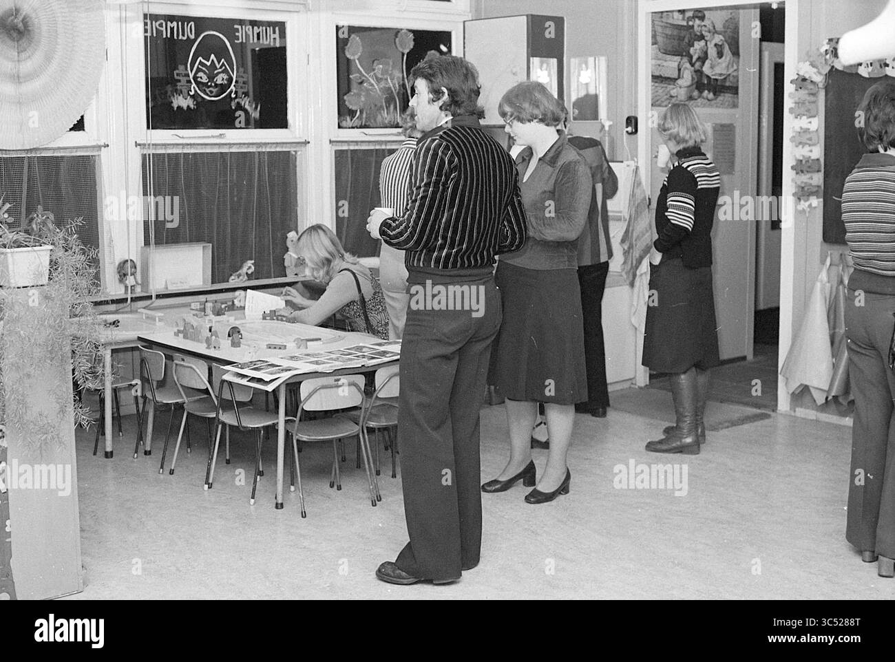 Open House Nursery Humpie Dumpie, IJm., open day e open House, 23-09-1976 Whizgle News, Dutch Desk, Paesi Bassi, 1950-2000 Un gruppo di persone è impegnato in varie attività in un ambiente intimo al coperto. Alcuni individui sono concentrati su un tavolo che mostra giochi o opere d'arte, mentre altri si trovano nelle vicinanze, chiacchierando o osservando. L'ambiente suggerisce un incontro informale, possibilmente in uno spazio comunitario o in un laboratorio creativo. Foto Stock