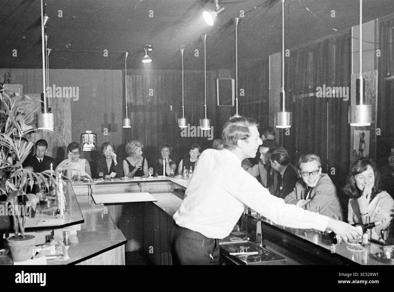 Alcuni signore e signori in un bar Whizgle News, Dutch Desk, Paesi Bassi, 1950-2000 Un vivace bar con clienti impegnati in conversazione mentre un barista prepara un drink dietro il bancone. L'illuminazione soffusa crea un'atmosfera intima, con un mix di ospiti elegantemente vestiti che si godono il loro tempo. Foto Stock