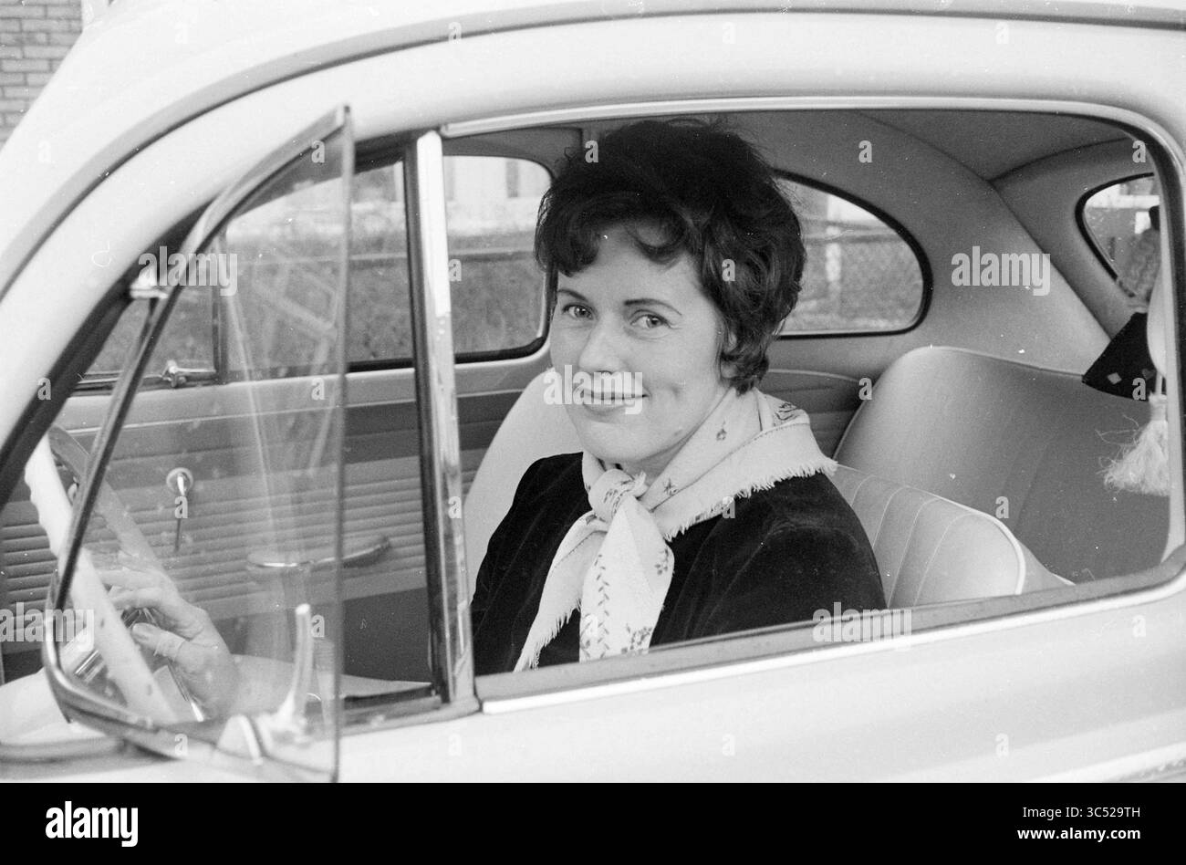 Donna al volante, 00-01-1962 Whizgle News, Dutch Desk, Paesi Bassi, 1950-2000 Una donna con un affascinante sorriso siede al posto di guida di un'auto d'epoca, vestita elegantemente con una sciarpa intorno al collo, trasuda un senso di nostalgia e stile classico. Foto Stock