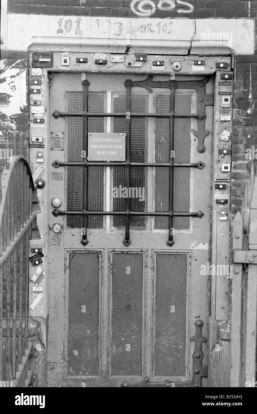 40 campanelli, collezioni, collezionisti, Amsterdam, Paesi Bassi, 10-04-1985 Whizgle News, Dutch Desk, Paesi Bassi, 1950-2000 una vecchia porta intemprata è adornata da una moltitudine di piccoli bottoni e interfoni, suggerendo un'entrata movimentata piena di storie. Un cartello apposto sulla porta aggiunge una nota di curiosità, invitante interazione, come suggerisce la storia dell'edificio. Il cancello circostante presenta intricati lavori in ferro, aggiungendo carattere alla scena. Foto Stock