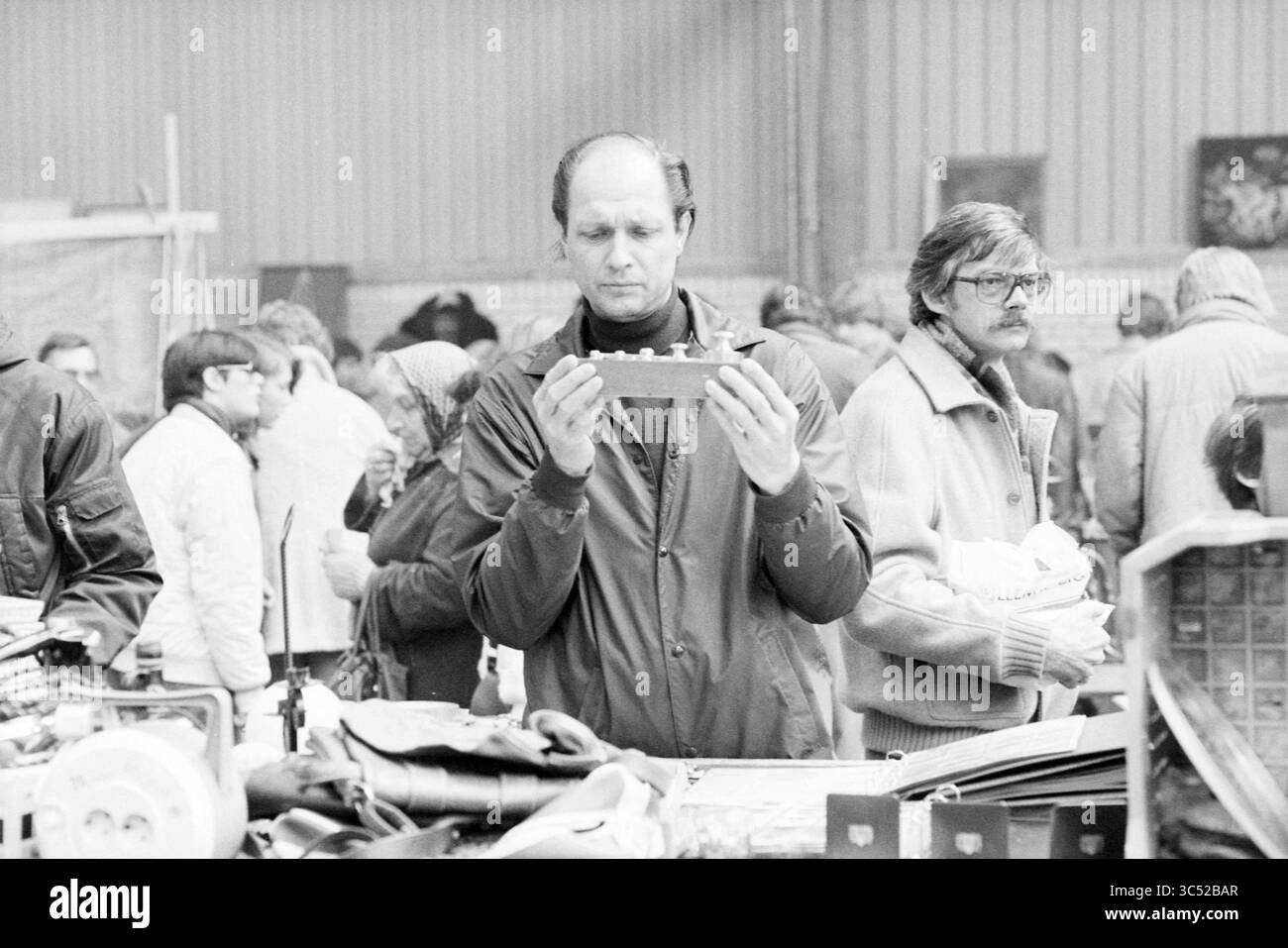 Report Black Market Beverwijk, Market, 00-00-1981 Whizgle News, Dutch Desk, Paesi Bassi, 1950-2000 Un uomo esamina con attenzione un piccolo oggetto nelle sue mani, circondato da una folla affollata di persone impegnate in varie attività in un mercato. Un altro individuo si trova nelle vicinanze, osservando la scena con un'espressione premurosa. Foto Stock