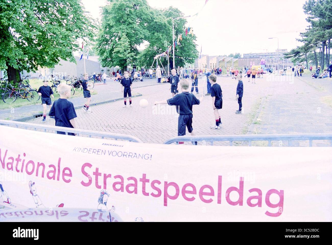 Street Play Day, Howler Buoy, IJmuiden, IJmuiden, Paesi Bassi, 23-05-2000 Whizgle News, Dutch Desk, Paesi Bassi, 1950-2000 Un vivace evento di gioco di strada con bambini impegnati in varie attività, con uno striscione che annuncia "Nationale Straatspeeldag" in evidenza. Le biciclette sono parcheggiate nelle vicinanze e gli alberi offrono ombra mentre gruppi di bambini partecipano a giochi e giochi. Foto Stock