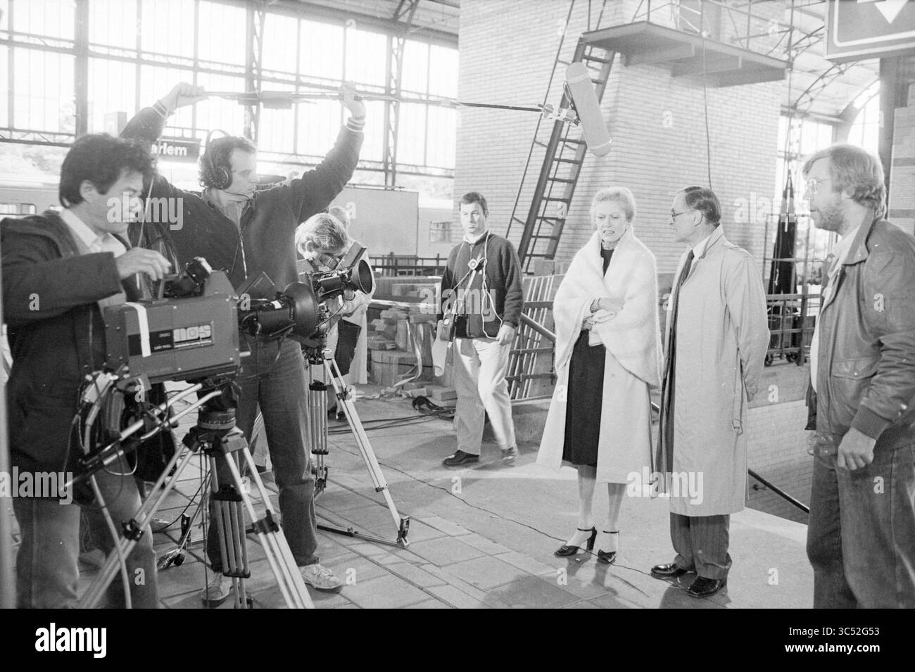 Registrazioni di film stazione di Haarlem, riprese, registrazioni di film, Haarlem, Stationsplein, paesi Bassi, 14-06-1984 Whizgle News, Dutch Desk, Paesi Bassi, 1950-2000 Una troupe cinematografica si prepara a girare in un grande spazio industriale, con telecamere e attrezzature pronte. In primo piano, un'attrice vestita elegantemente è accanto a un regista o produttore, mentre i membri della troupe regolano la loro posizione e la loro attrezzatura. L'atmosfera suggerisce una giornata intensa di riprese, con i preparativi in corso per una scena importante. Foto Stock