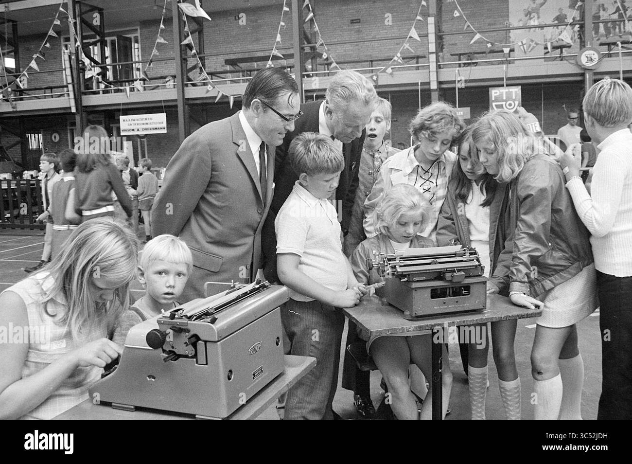 Fiera del lavoro per giovani, 00-07-1970 Whizgle News, Dutch Desk, Paesi Bassi, 1950-2000 Un gruppo di adulti e bambini si riuniscono intorno a un tavolo, esaminando curiosamente le macchine da scrivere mentre si impegnano in una conversazione animata. Alcuni bambini sono concentrati sul funzionamento delle macchine, mentre altri guardano con attenzione, creando un'atmosfera vivace. Lo sfondo presenta decorazioni e un accenno di un evento vivace. Foto Stock