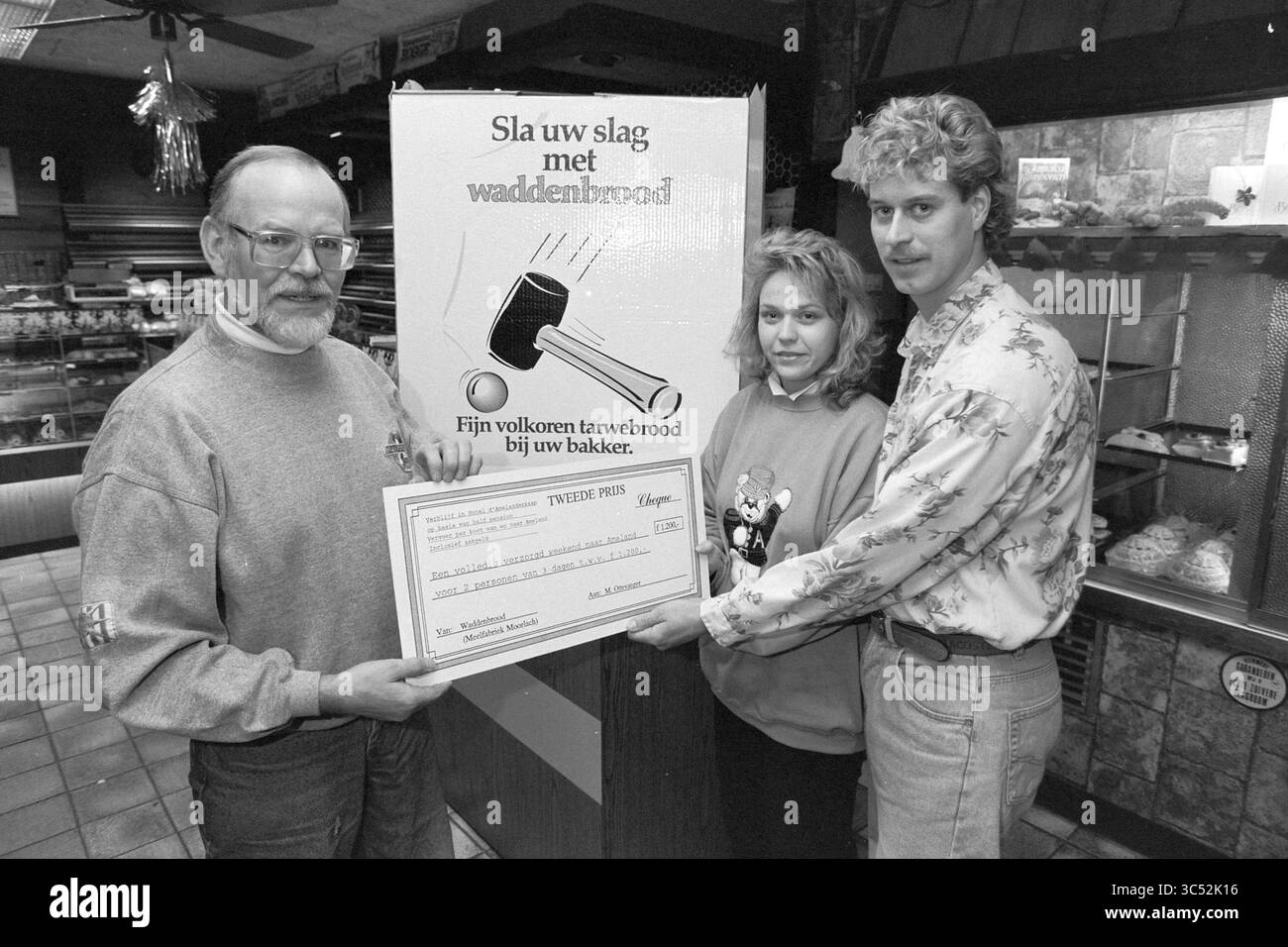 Cerimonia di premiazione per baker Verkerk, 11-12-1991 Whizgle News, Dutch Desk, Paesi Bassi, 1950-2000 tre persone stanno insieme in un ambiente accogliente e rustico, tenendo con orgoglio un grande assegno. Un cartello dietro di loro promuove il "waddenbrood", un tipo di pane integrale, ed esprime il sostegno ai panettieri locali. Ogni persona indossa un abbigliamento casual e le sue espressioni trasmettono un senso di realizzazione e spirito comunitario. Foto Stock