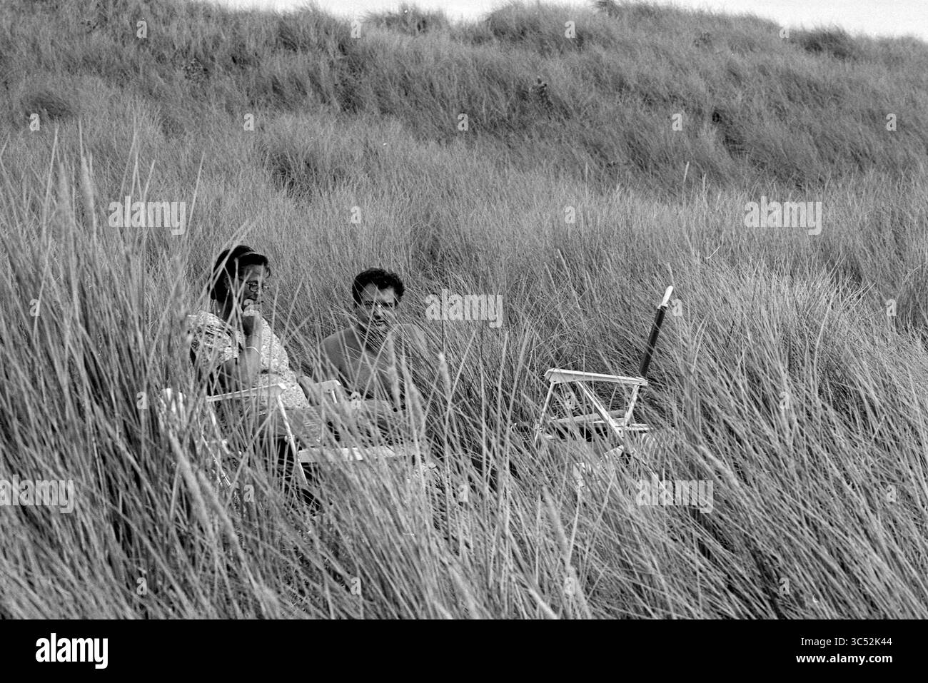 Nuove dune IJmuiderslag, 17-08-1993 Whizgle News, Dutch Desk, Paesi Bassi, 1950-2000 due persone siedono tra erba alta, impegnati in conversazione. Sembrano rilassate, circondate dalla natura, mentre godono di un momento di pace insieme. Una sedia e un cavalletto suggeriscono la presenza di attività artistiche o di svago. Foto Stock