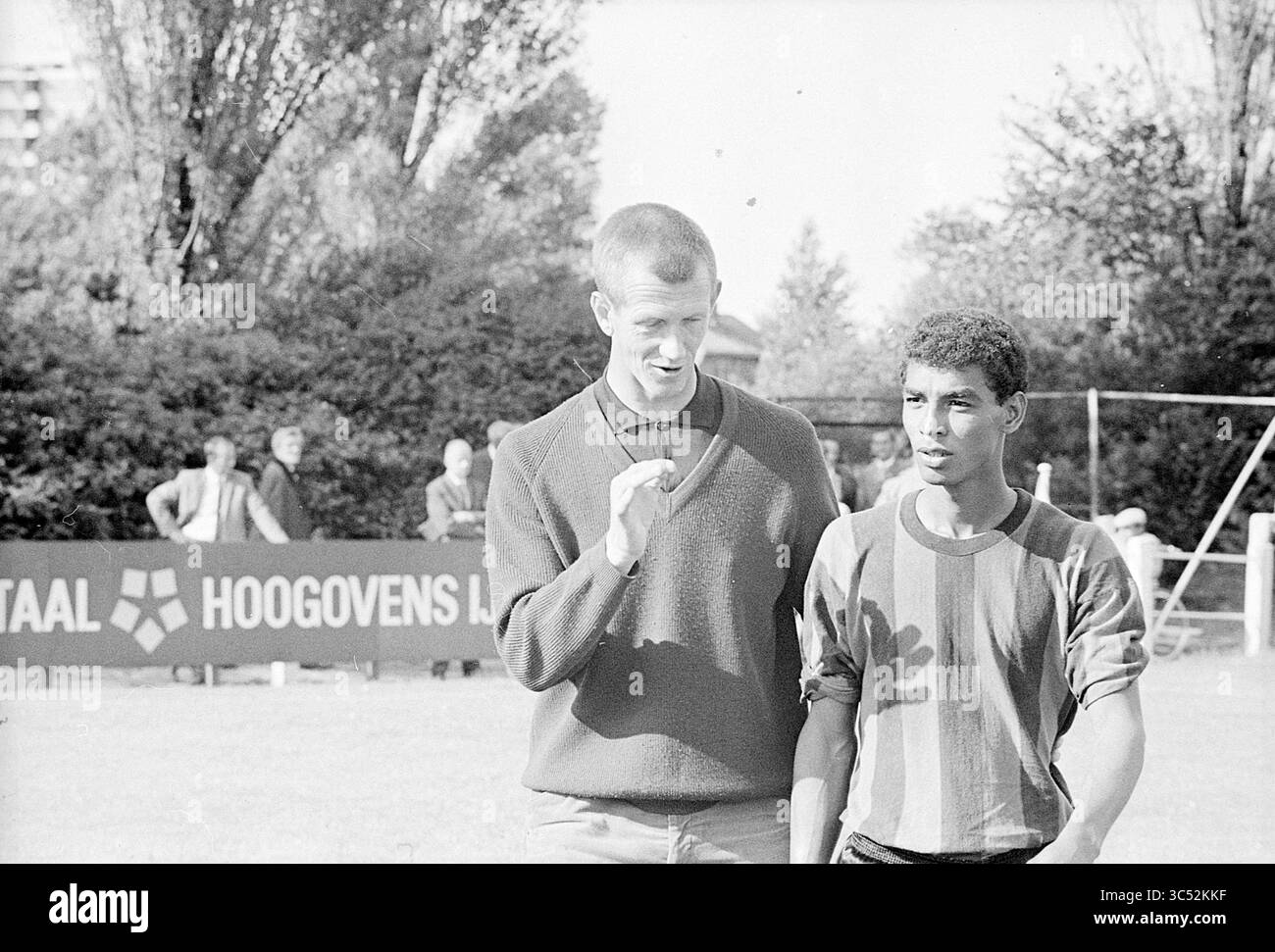 Calcio maschile Whizgle News, Dutch Desk, Paesi Bassi, 1950-2000 due persone si impegnano in conversazione, con un gesto, mentre l'altro ascolta con attenzione. Sembrano essere all'aperto, circondati dal verde e da un raduno di persone sullo sfondo, suggerendo un'atmosfera vivace. Foto Stock