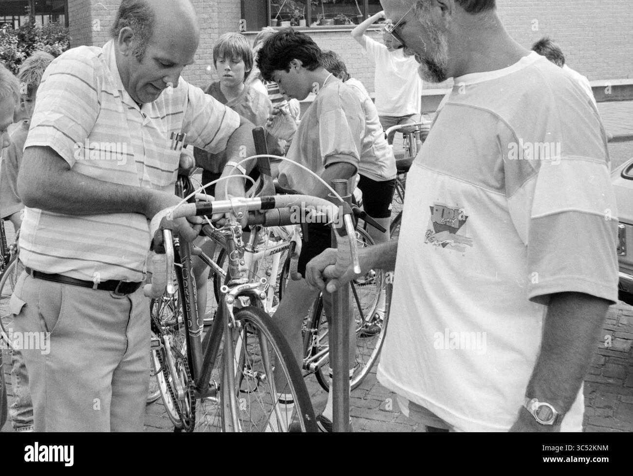 Ispezione biciclette Mosquito Round, Mosquito Round, 10-08-1988 Whizgle News, Dutch Desk, Paesi Bassi, 1950-2000 due uomini stanno lavorando insieme su una bicicletta, ispezionando il suo manubrio e altri componenti. Sullo sfondo, un gruppo di giovani osserva l'attività, mostrando interesse per il processo di riparazione della moto. Foto Stock