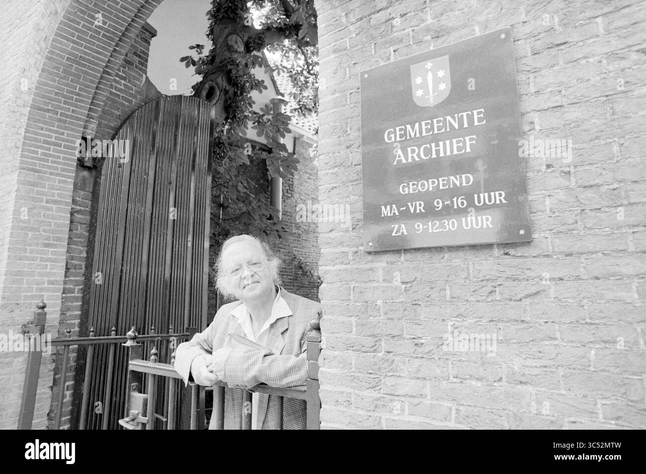 Kulker at Haarlem Municipal Archives, Haarlem, Jansstraat, Paesi Bassi, 24-06-1992 Whizgle News, Dutch Desk, Paesi Bassi, 1950-2000 un individuo anziano si trova accanto a un cancello, appoggiato al muro con un'espressione accogliente. Un cartello nelle vicinanze indica gli orari di apertura dell'archivio municipale locale, evidenziando il suo ruolo nel preservare la storia della comunità. Texture di pietra e vegetazione strisciante aggiungono un tocco di atmosfera alla scena. Foto Stock