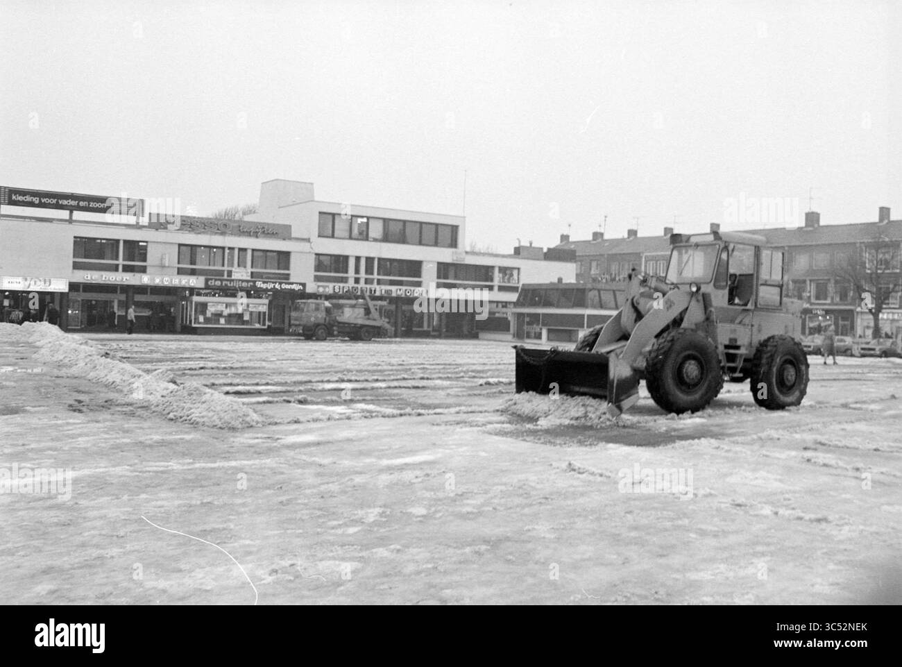 Disinnevamento da Marktplein IJmuiden, IJmuiden, Marktplein, Paesi Bassi, 30-01-1979 Whizgle News, Dutch Desk, Paesi Bassi, 1950-2000 Una macchina per impieghi gravosi sgombra la neve da una piazza deserta della città, rivelando aree di pavimentazione mentre edifici moderni si trovano sullo sfondo. La scena enfatizza l'atmosfera fredda e invernale e l'impatto del clima invernale sulla vita urbana. Foto Stock