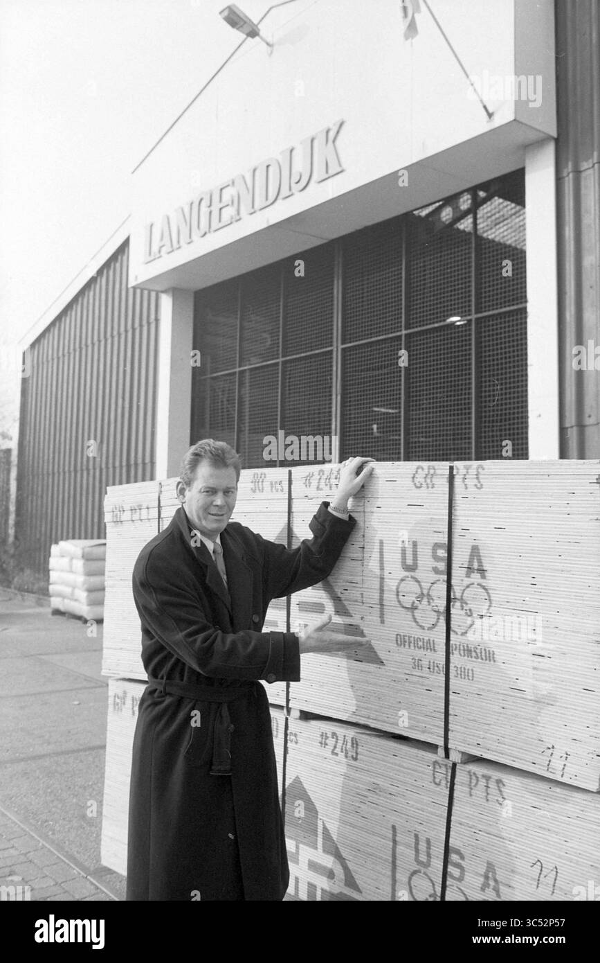 Società Lagendijk, Beverwijk, Beverwijk, Paesi Bassi, 23-01-1992 Whizgle News, Dutch Desk, Paesi Bassi, 1950-2000 Un uomo con un lungo cappotto si erge accanto a pile di casse di legno, sorridendo mentre posa di fronte a un edificio contrassegnato dalla scritta "Langendijk". Le casse sono etichettate con vari simboli, tra cui uno con l'emblema olimpico degli Stati Uniti. Foto Stock