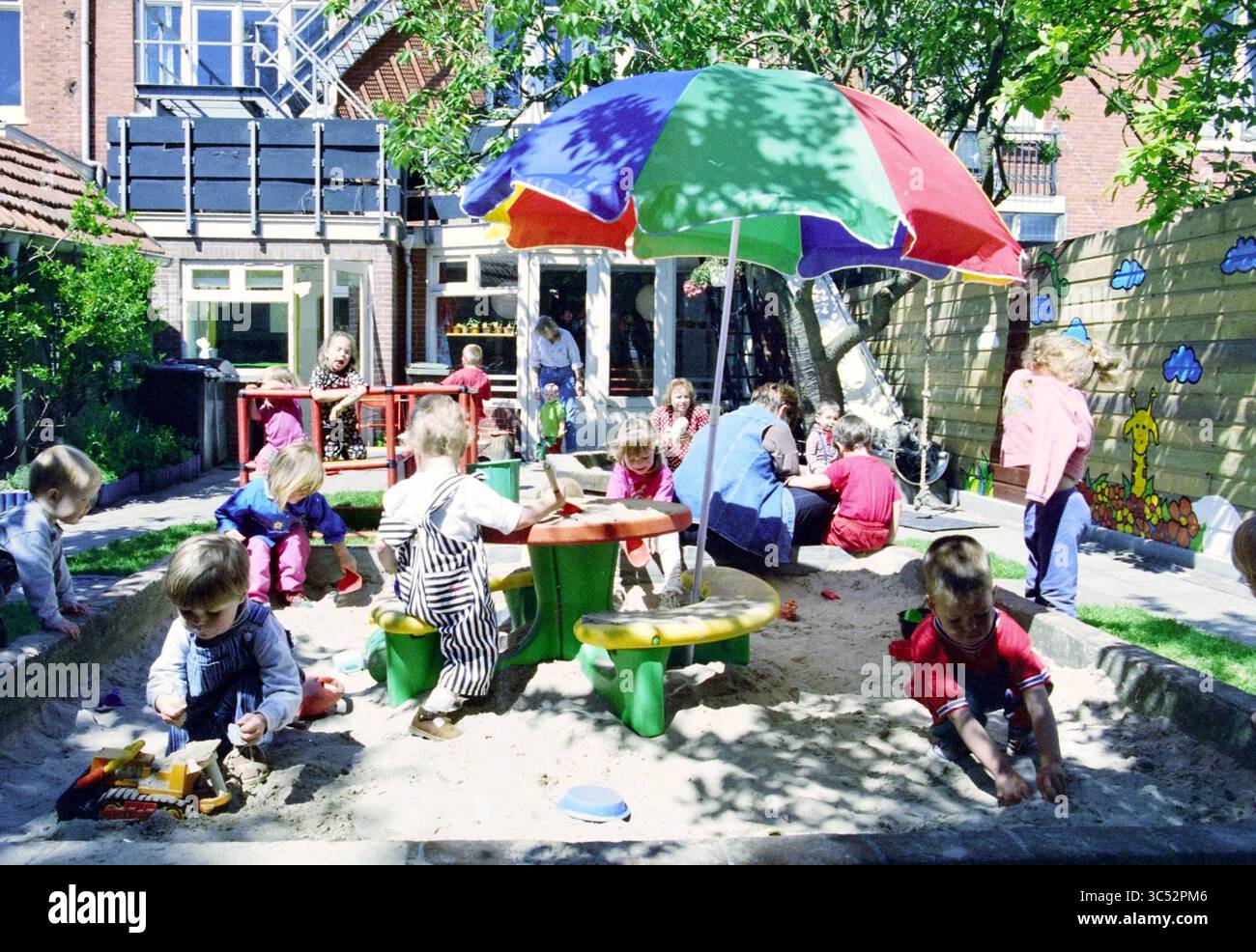 Garden with Children, Pimpernel, 18-06-1995 Whizgle News, Dutch Desk, Paesi Bassi, 1950-2000 Un vivace parco giochi pieno di bambini impegnati in varie attività, tra cui giocare in un colorato sandbox sotto un ombrello vivace, mentre altri interagiscono ed esplorano intorno a loro. L'atmosfera è allegra e giocosa, con adulti che supervisionano nelle vicinanze. Foto Stock