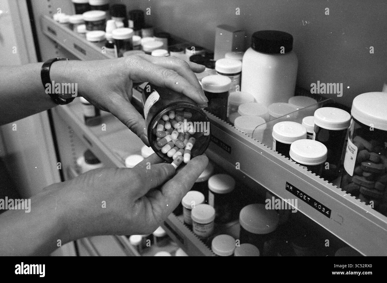 Donna prende barattolo di capsule dal gabinetto di medicina, 22-12-1982 Whizgle News, Dutch Desk, Paesi Bassi, 1950-2000 Una persona sta versando attentamente le capsule da una bottiglia di prescrizione, circondato da vari contenitori di farmaci ordinatamente disposti sugli scaffali. Foto Stock
