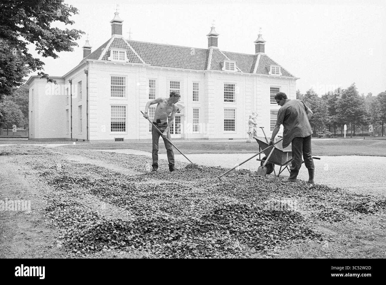 Giardinieri in azione di fronte alla casa di campagna di Beeckestijn Whizgle News, Dutch Desk, Paesi Bassi, 1950-2000 due uomini lavorano all'aperto, rastrellando e spargendo ghiaia in un'area giardino di fronte a un grande e maestoso edificio dall'architettura tradizionale. Una carriola si trova nelle vicinanze, pronta ad assistervi con il loro compito. La scena riflette lo sforzo di mantenere i giardini, evidenziando una miscela di duro lavoro e fascino storico. Foto Stock