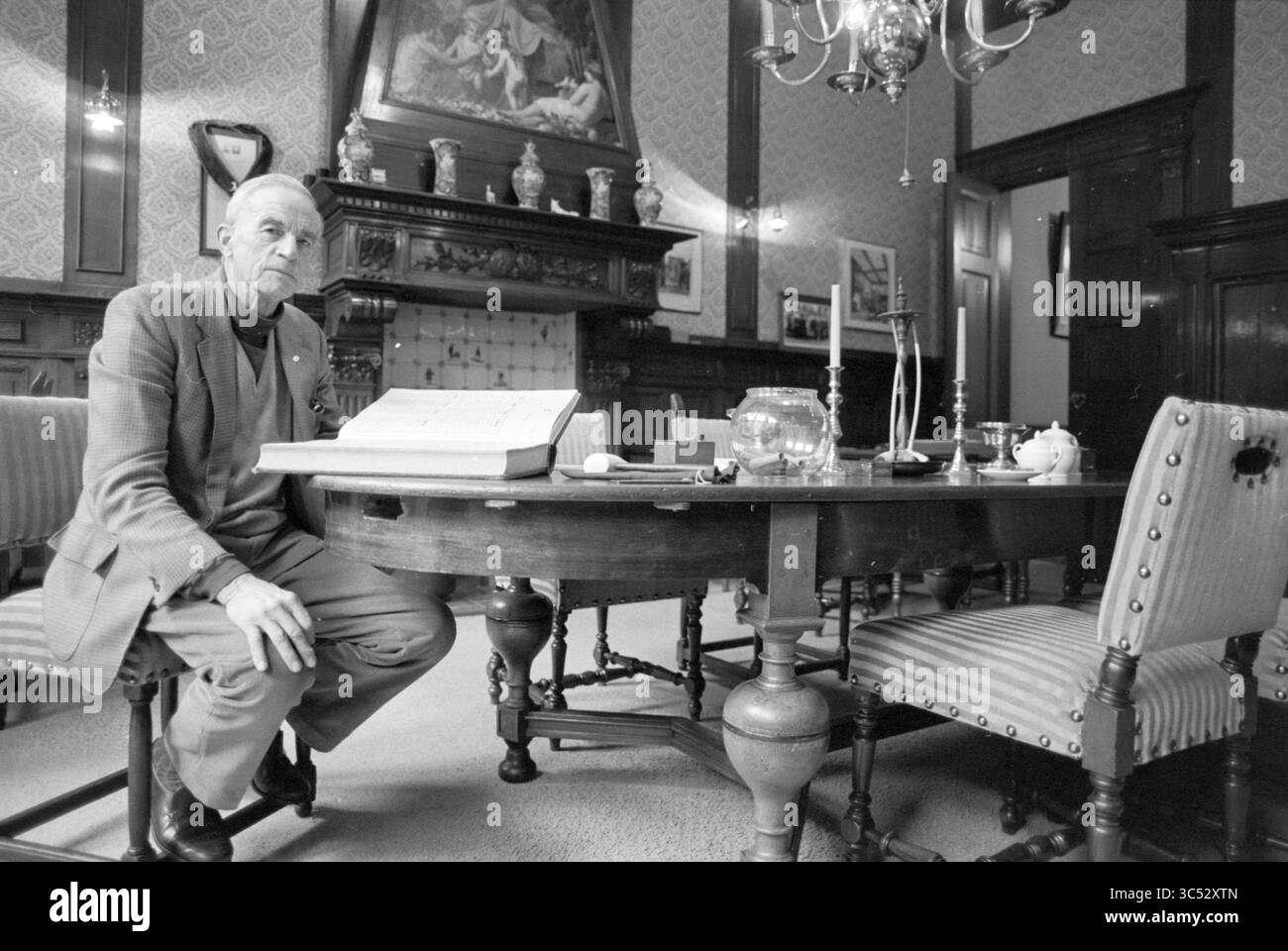 Uomo in una stanza d'antiquariato Whizgle News, Dutch Desk, Paesi Bassi, 1950-2000 un anziano gentiluomo seduto ad un tavolo da pranzo in legno in una sala riccamente decorata. Circondato da arredi eleganti e da un decor sottile, appare profondamente nel pensiero, con un libro aperto che riposa davanti a lui. La camera presenta dettagli ornamentali, tra cui un lampadario e un grande dipinto sulla parete, che aggiungono al suo fascino classico. Foto Stock