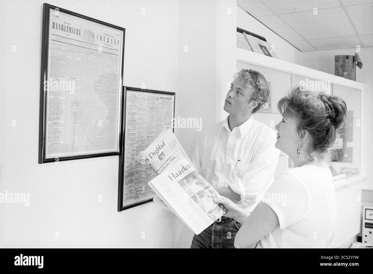 Guardando i vecchi giornali, Haarlems Dagblad, 12-07-1995 Whizgle News, Dutch Desk, Paesi Bassi, 1950-2000 due persone esaminano un articolo di giornale storico incorniciato su un muro, mentre uno tiene un altro giornale. Le loro espressioni trasmettono curiosità e coinvolgimento con i contenuti visualizzati. Foto Stock