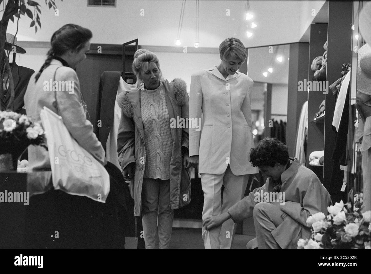 El Rose, Walk-in show, 08-03-1994 Whizgle News, Dutch Desk, Paesi Bassi, 1950-2000 Un gruppo di quattro persone viene riunito in un negozio di abbigliamento, con una persona che prova un abito elegante e personalizzato. Due donne stanno osservando e offrendo feedback, mentre un individuo più giovane si inginocchia per regolare l'abbigliamento. L'ambiente è pieno di vari indumenti e accessori, creando un'atmosfera vivace incentrata sulla moda e lo stile. Foto Stock