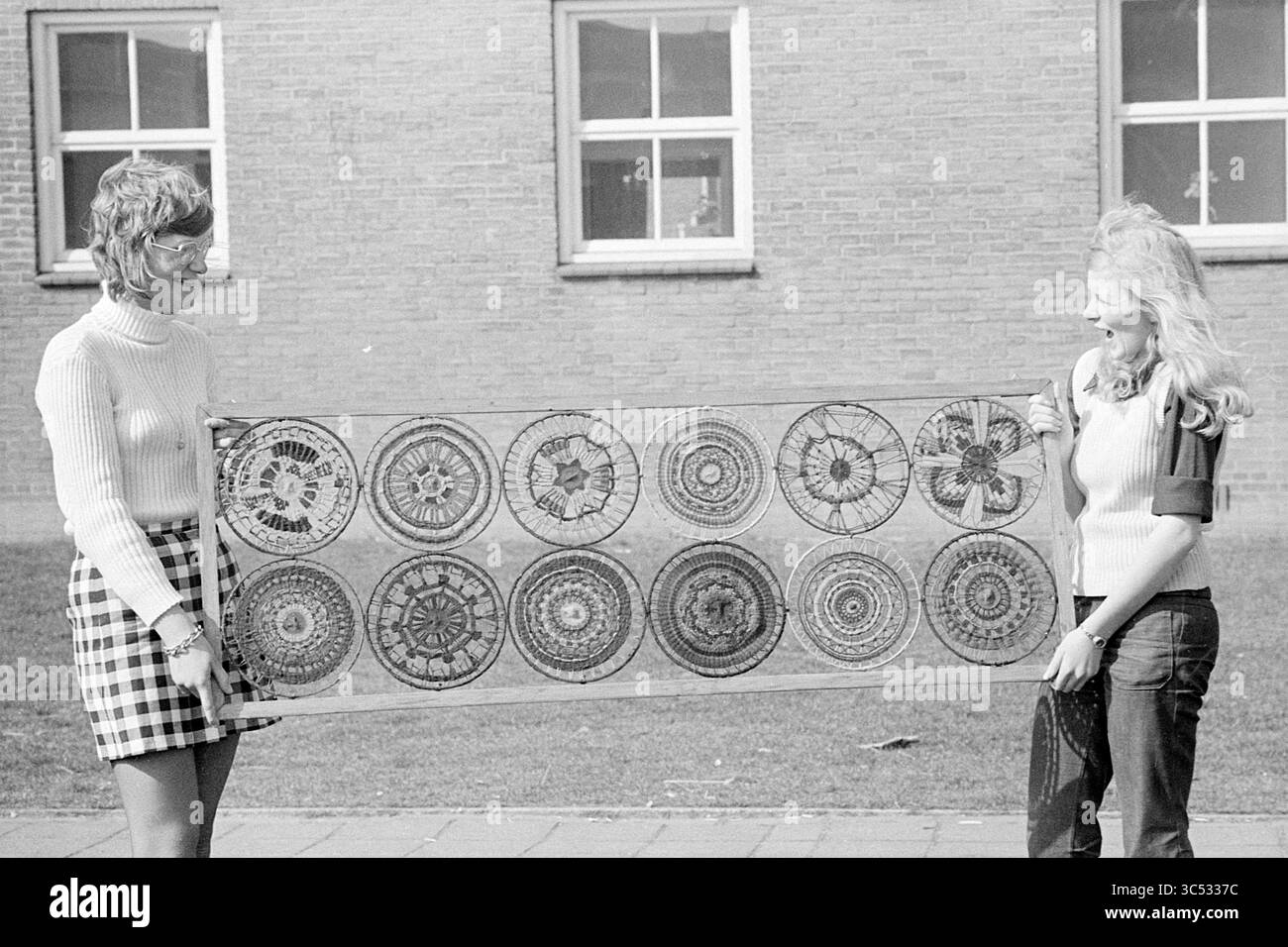 Ragazze con opere d'arte. Patchwork o uncinetto. Whizgle News, Dutch Desk, Paesi Bassi, 1950-2000 due donne stanno all'aperto, tenendo un lungo tessuto con intricati motivi circolari. Sono realizzati in stile retrò, con uno che indossa un pullover a coste e l'altro un top elegante con una gonna a quadri. Lo sfondo presenta un edificio in mattoni e finestre. Foto Stock