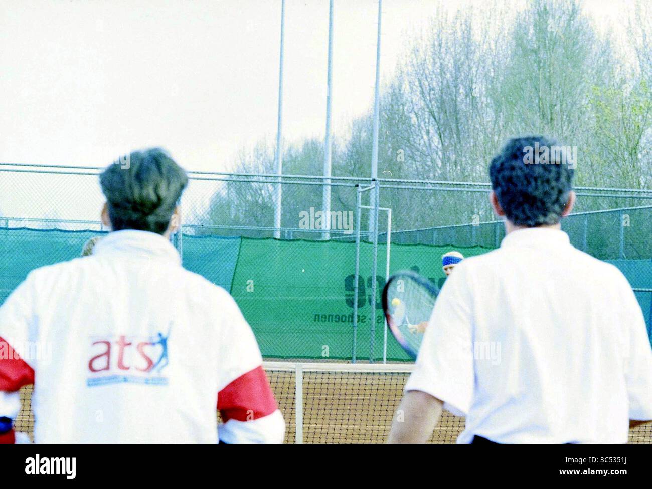 Apertura della stagione di tennis Arnoldus Park, 03-04-1995 Whizgle News, Dutch Desk, Paesi Bassi, 1950-2000 due persone in abbigliamento atletico guardano intensamente come una partita di tennis si svolge in lontananza, con i giocatori impegnati su un campo circondato da una recinzione e alberi sullo sfondo. Foto Stock