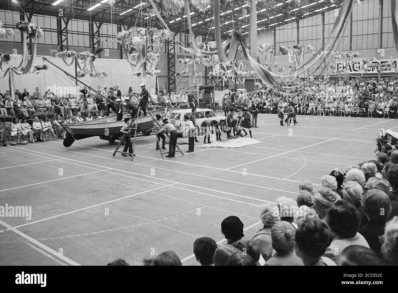 Addio burg. Cremers Sports Hall, Mayors, 30-05-1969 Whizgle News, Dutch Desk, Paesi Bassi, 1950-2000 Un vivace evento al coperto presenta uno spettacolo con un'auto e una barca sul palco, circondati da spettatori entusiasti. Decorata con striscioni e streamer natalizi, la scena cattura un momento di intrattenimento e coinvolgimento della comunità, con artisti impegnati in attività che enfatizzano il lavoro di squadra e le abilità. Il pubblico, pieno di una folla diversificata, guarda attentamente, aggiungendo un'atmosfera vivace. Foto Stock