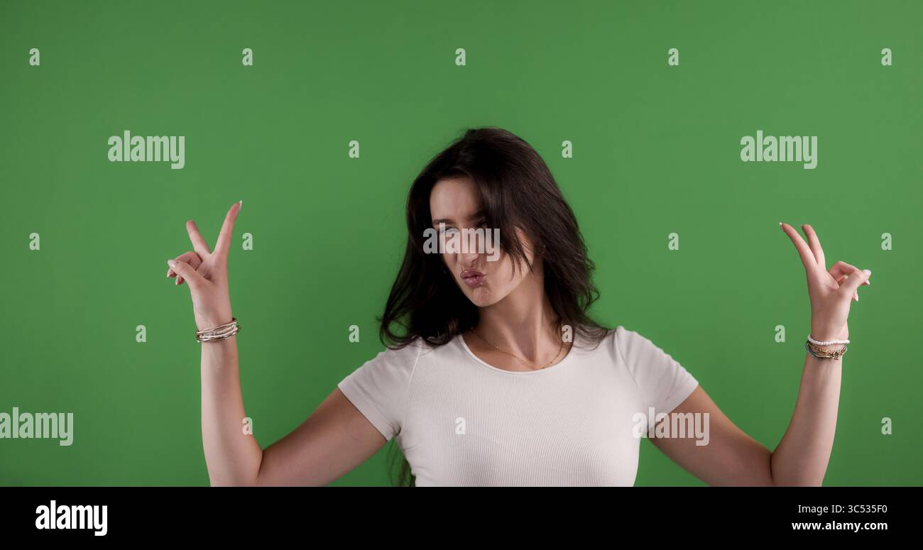 Giovane donna che fa un volto divertente e mostra un segno di pace con entrambe le mani sullo sfondo della chiave cromatica Foto Stock
