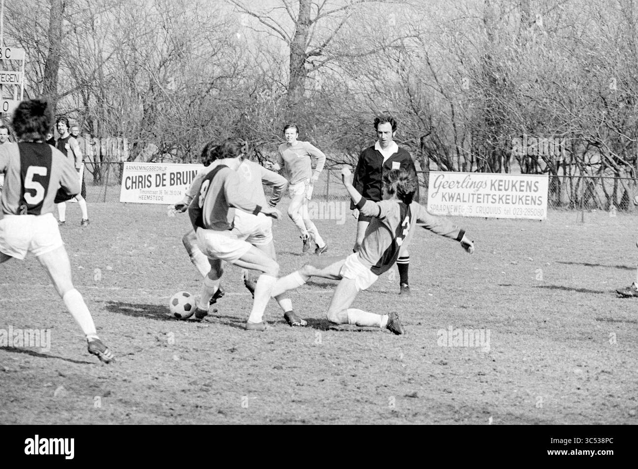 HBC - Fulmars, Football, 28-03-1976 Whizgle News, Dutch Desk, Paesi Bassi, 1950-2000 Una partita di calcio dinamica si svolge su un campo erboso, dove i giocatori sono impegnati in un gioco intenso. Un giocatore difende abilmente contro un avversario, che è a terra dopo un placcaggio. La scena cattura l'emozione del gioco, con spettatori e banner pubblicitari sullo sfondo. Foto Stock