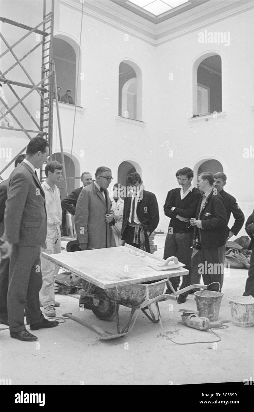 L'intonacatore spiega agli scolari (possibilmente inglese), 00-00-1966 Whizgle News, Dutch Desk, Paesi Bassi, 1950-2000 Un gruppo di uomini e giovani studenti si riuniscono intorno a una carriola piena di materiali da costruzione, impegnati in una discussione all'interno di un edificio spazioso e infinito con finestre ad arco. Ponteggi e utensili sono presenti, a indicare un progetto di ristrutturazione o costruzione attivo. Foto Stock