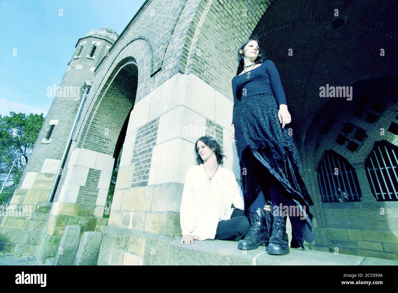 Family Van Rossum, Haarlem, Paesi Bassi, 24-09-2002 Whizgle News, Dutch Desk, i Paesi Bassi, 1950-2000 due persone si alzano e si siedono vicino a un grande arco testurizzato, vestiti con stili contrastanti che mescolano eleganza ed eleganza. Uno si regge con sicurezza con una gonna lunga e fluida e stivali, mentre l'altro si siede con grazia, indossando un outfit più leggero. L'atmosfera è artistica e leggermente stravagante, sullo sfondo della struttura architettonica. Foto Stock