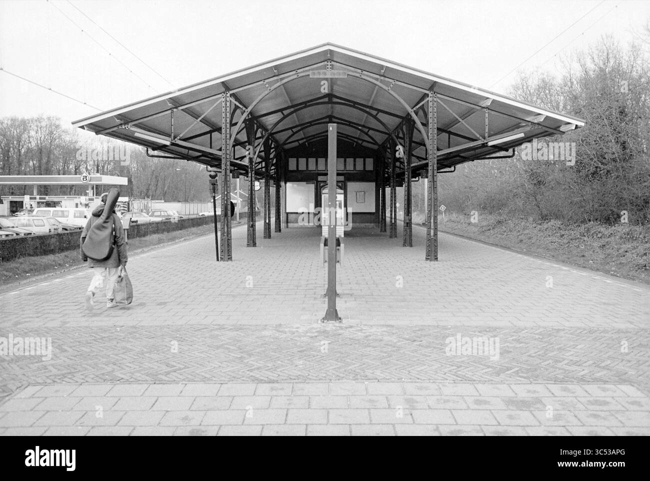 Panoramica della piattaforma presso La stazione S'poort Zuid, Santpoort-Zuid, 11-12-1994 Whizgle News, Dutch Desk, Paesi Bassi, 1950-2000 Una figura solitaria cammina lungo una piattaforma deserta, caratterizzata da un baldacchino dal design sorprendente supportato da intricati lavori in ferro. L'area circostante è tranquilla, con un pizzico di natura che si insinua dai bordi, creando un senso di quiete e attesa. Foto Stock