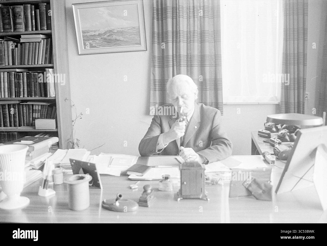 Notaio Boelaagen dietro la scrivania, persone, 22-03-1971 Whizgle News, Dutch Desk, Paesi Bassi, 1950-2000 un anziano gentiluomo si siede su una scrivania disordinata, nel profondo del pensiero mentre esamina un documento. Dietro di lui, scaffali pieni di libri fiancheggiano il muro, e una finestra lascia entrare luce soffusa, mettendo in risalto l'atmosfera serena dell'ufficio. Un assortimento di articoli da ufficio e di articoli personali è sparso su tutta la scrivania, aggiungendo la sensazione di uno spazio di lavoro occupato ma contemplativo. Foto Stock