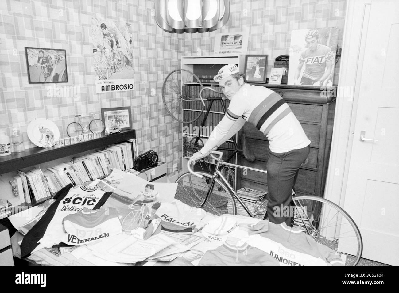 Piet Hoogland, collezionista di ciclismo, Haarlem, persone, collezioni, collezionisti, Haarlem, Paesi Bassi, 24-12-1980 Whizgle News, Dutch Desk, i Paesi Bassi, 1950-2000 Un ciclista posa in una stanza piena di cimeli del ciclismo d'epoca, tra cui maglie e poster, mentre regola una bicicletta accanto a una mostra ben organizzata di attrezzatura da ciclismo. L'arredamento presenta motivi classici e tocchi nostalgici, che riflettono la passione per lo sport. Foto Stock