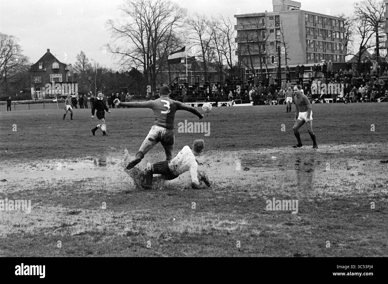 Partita di calcio Royal HFC su un campo bagnato immerso possibile contro Old Internationals Whizgle News, Dutch Desk, Paesi Bassi, 1950-2000 Una partita di calcio fangosa si svolge mentre i giocatori navigano su un campo bagnato, con un giocatore che scivola in un tackle mentre un altro si prepara a dribblare. Gli spettatori guardano da lontano e gli edifici incorniciano la scena sullo sfondo, catturando l'intensità e le sfide del gioco in condizioni avverse. Foto Stock