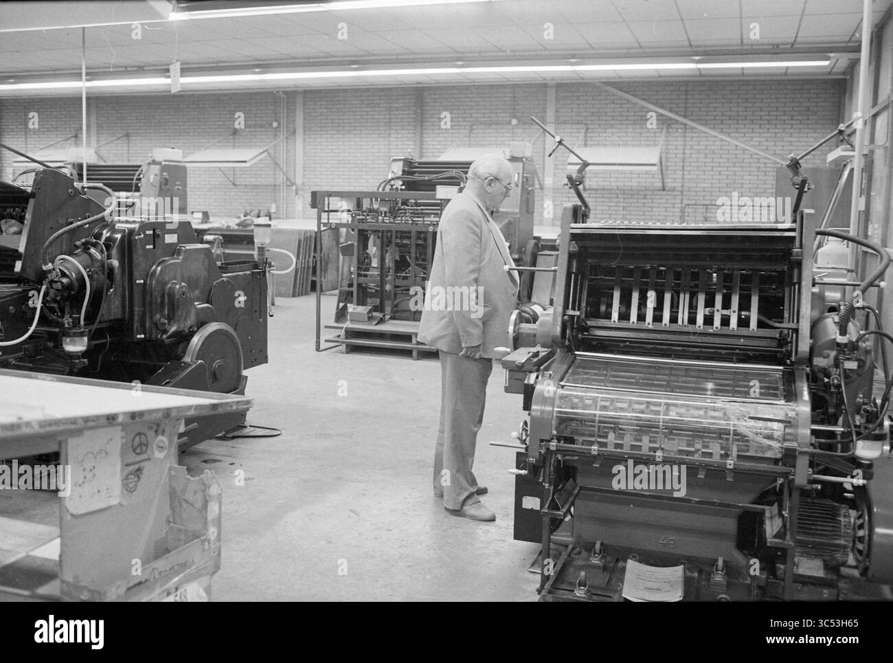 Asta per la stampa diurna Green IJmuiden, Auction, IJmuiden, Nederland, 03-11-1986 Whizgle News, Dutch Desk, Paesi Bassi, 1950-2000 Un gentiluomo osserva una macchina da stampa complessa in un'officina industriale, circondata da vari macchinari che suggeriscono una vivace attività di stampa. Lo spazio di lavoro presenta un assortimento di apparecchiature di stampa, evidenziando le complessità del processo di stampa. Foto Stock