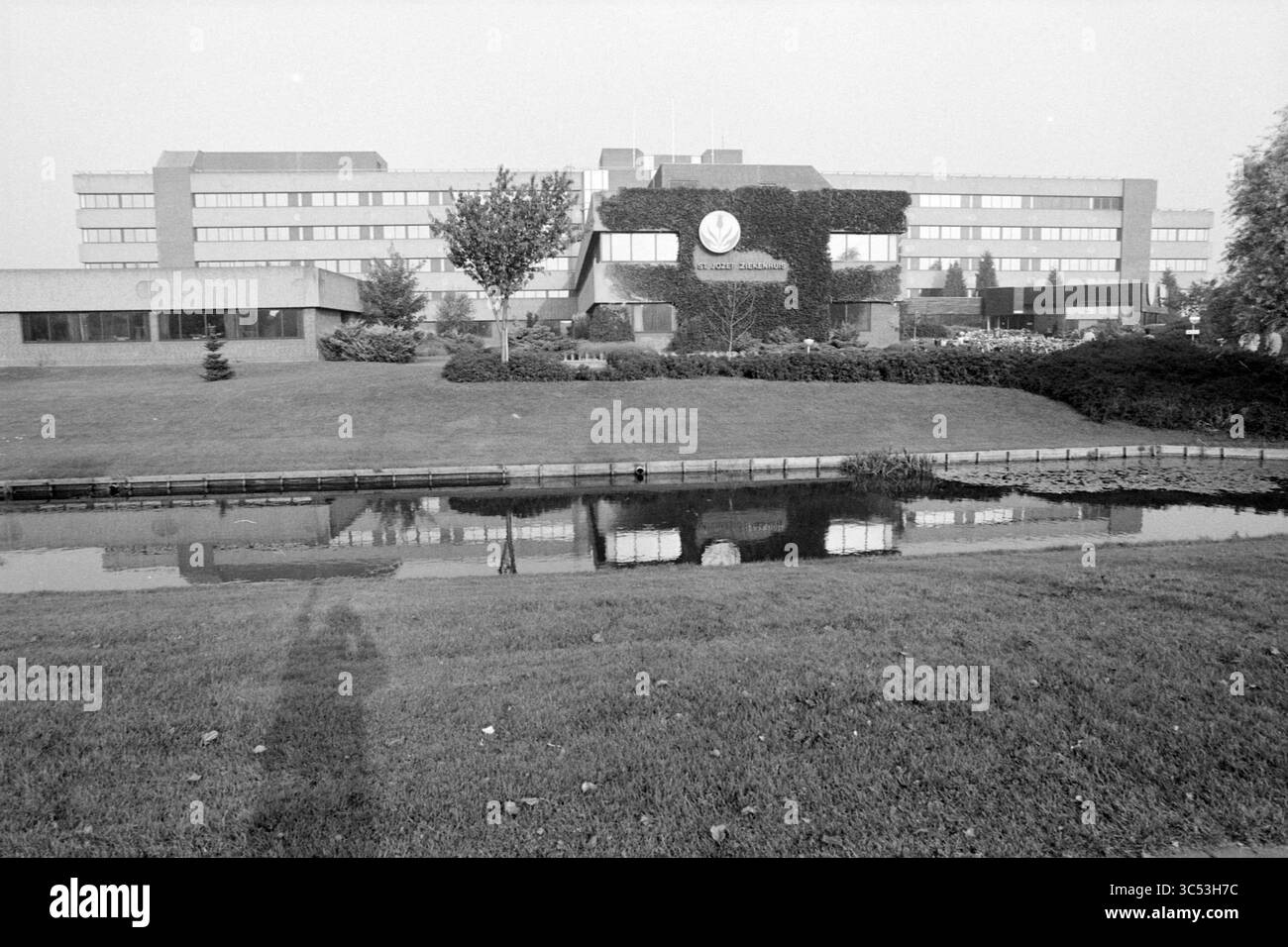 Hospital St. Jozef Heemskerk, ospedali, Heemskerk, 18-10-1989 Whizgle News, Dutch Desk, i Paesi Bassi, 1950-2000 Un edificio moderno con un prato frontale paesaggistico si riflette in un tranquillo specchio d'acqua nelle vicinanze. Il verde lussureggiante circonda la struttura, creando un'atmosfera serena. Il design presenta un emblema prominente, che aggiunge al carattere della struttura. Foto Stock