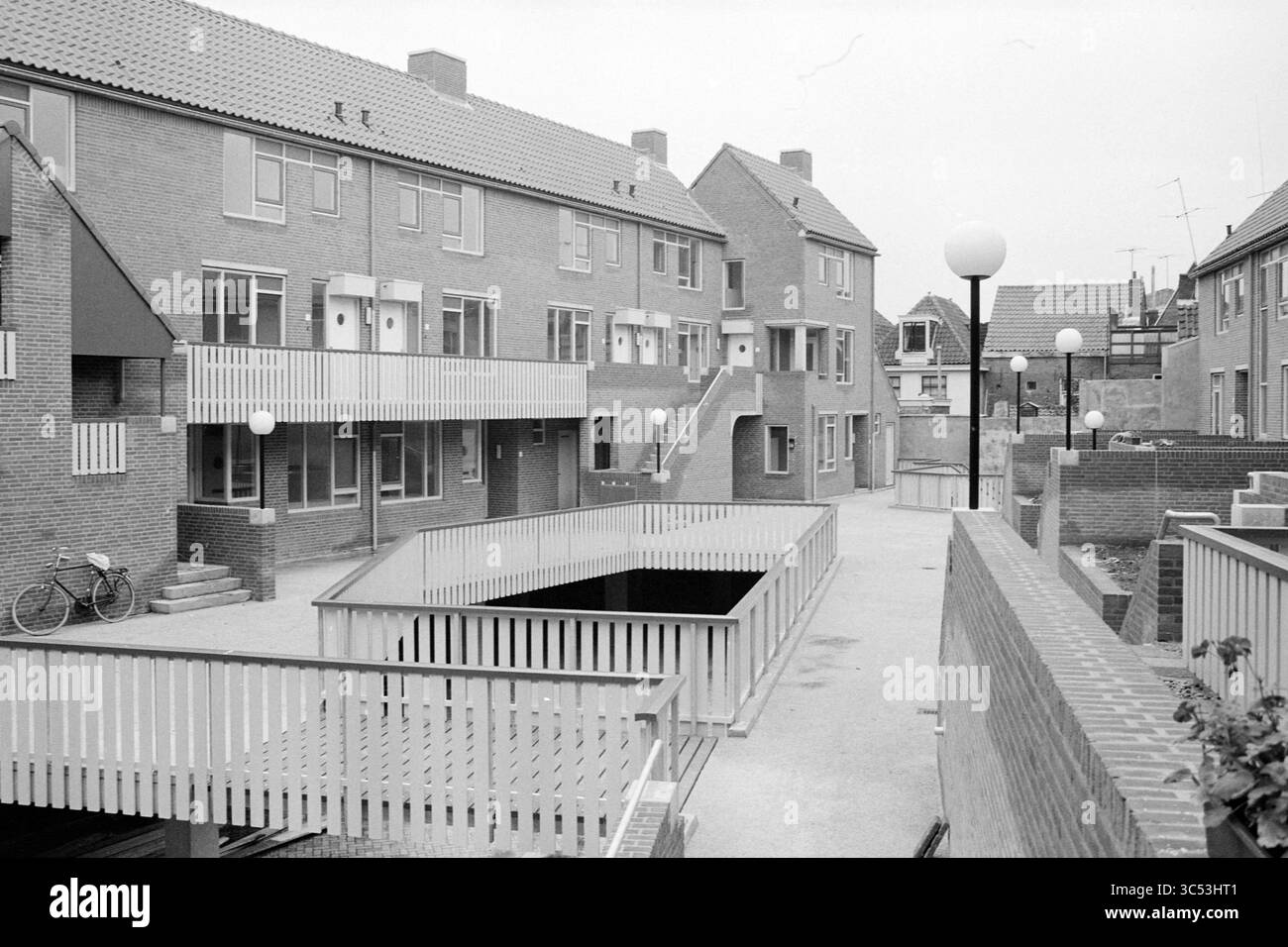 New Houses Butter Market Wall, Houses and House Construction, Haarlem, Botermarkt, Nederland, 11-10-1982 Whizgle News, Dutch Desk, Paesi Bassi, 1950-2000 Un pittoresco complesso residenziale caratterizzato da edifici in mattoni a due piani, interconnessi da passaggi pedonali e circondati da aree paesaggistiche. La disposizione comprende spazi esterni con deposito biciclette e lampade rotonde, creando un'atmosfera accogliente. Foto Stock