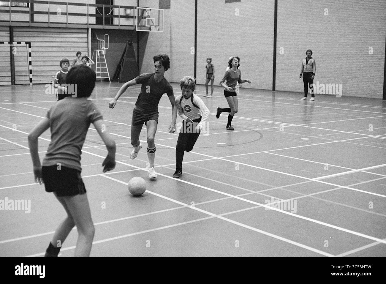 I bambini giocano a sport in una sala sportiva, 00-08-1977 Whizgle News, Dutch Desk, Paesi Bassi, 1950-2000 Una vivace partita di calcio al coperto presenta diversi bambini impegnati in un gioco vivace, con un ragazzo che manovra abilmente la palla mentre altri lo inseguono, mostrando il lavoro di squadra e l'eccitazione in una palestra scolastica. Foto Stock