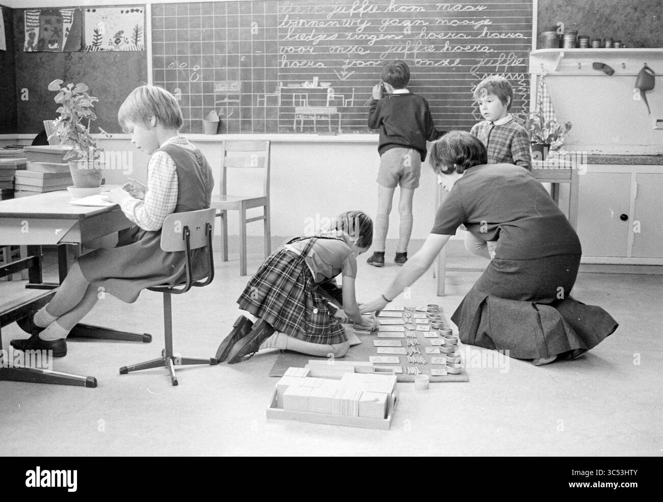 I bambini in una classe preparano qualcosa per il loro insegnante Whizgle News, Dutch Desk, Paesi Bassi, 1950-2000 Una scena in classe raffigurante bambini impegnati in varie attività. Un bambino si concentra sulla scrittura a una scrivania, mentre un altro si inginocchia per sistemare i materiali sul pavimento. Un insegnante sta organizzando gli oggetti nelle vicinanze, e altri due studenti sono in piedi su una lavagna coperta di scrittura. L'ambiente appare educativo e collaborativo, favorendo l'apprendimento e l'interazione tra gli studenti. Foto Stock