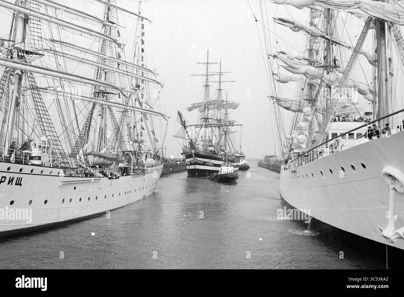 Vela: Chiuse nelle chiuse, 09-08-1990 Whizgle News, Dutch Desk, Paesi Bassi, 1950-2000 Una flotta di maestose navi alte naviga in uno stretto canale, con i loro alberi che torreggiano contro lo skyline. Le navi sono adornate di sartiame e vele, mentre le imbarcazioni più piccole aiutano a manovrare in mezzo alla movimentata attività del porto. Foto Stock