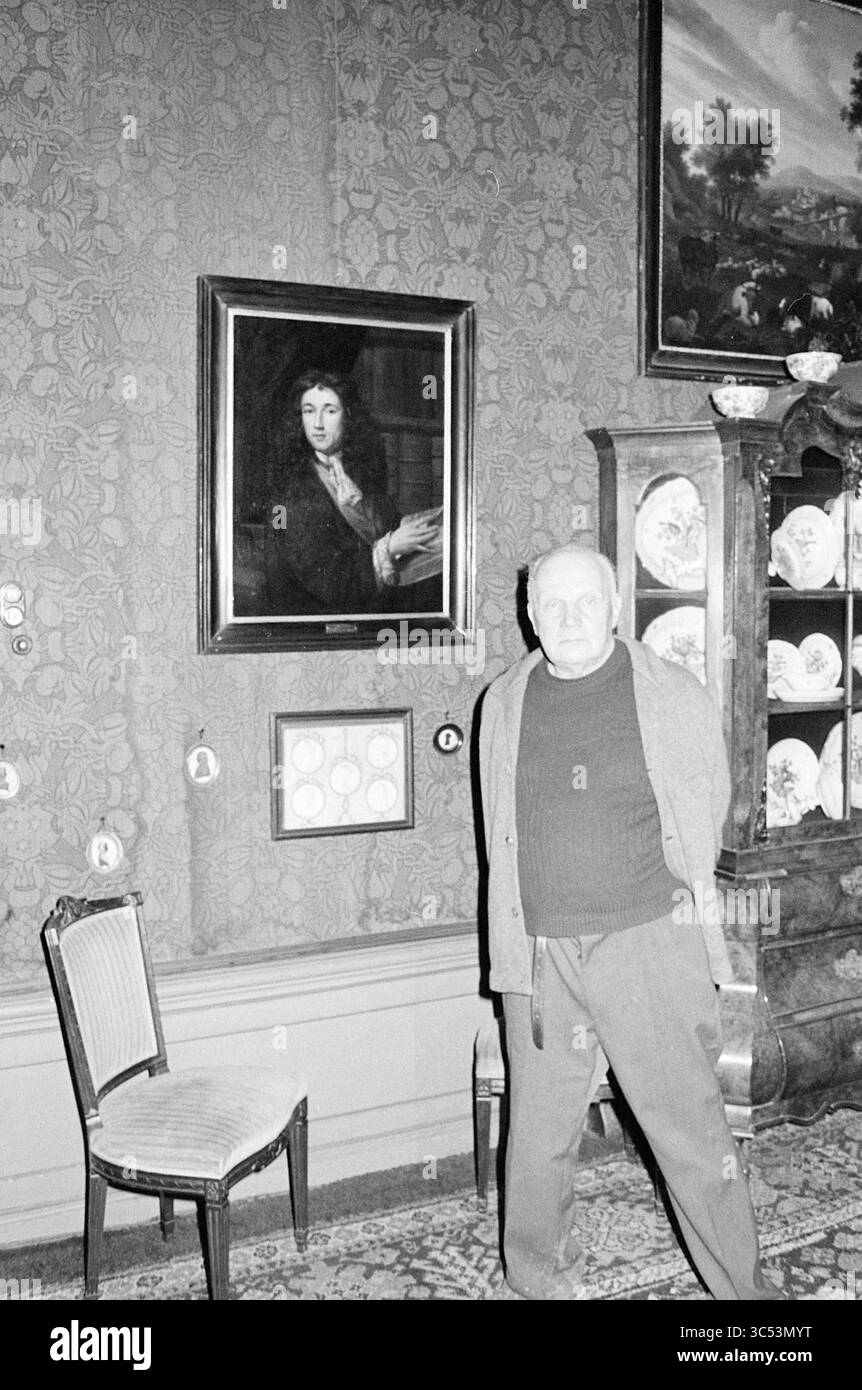 Baron by Ghost painting, Ghosts, 04-12-1977 Whizgle News, Dutch Desk, Paesi Bassi, 1950-2000 un uomo anziano si trova vicino a un grande ritratto incorniciato appeso su una parete a motivi geometrici, circondato da lastre decorative esposte in un armadietto. Una sedia di legno è posizionata nelle vicinanze, a complemento dell'atmosfera storica della camera. Foto Stock
