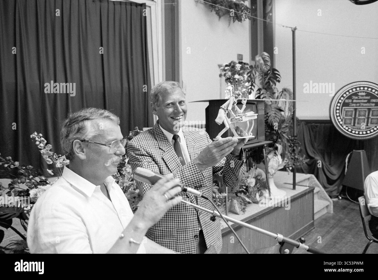 Addio Theo Braak, voce & VR., 06-07-1991 Whizgle News, Dutch Desk, Paesi Bassi, 1950-2000 due uomini si trovano davanti a uno sfondo festoso, con uno che tiene in alto un trofeo, mentre l'altro parla in un microfono. Intorno a loro si trovano piante decorative e un emblema circolare in mostra, che si aggiunge all'atmosfera celebrativa dell'evento. Foto Stock