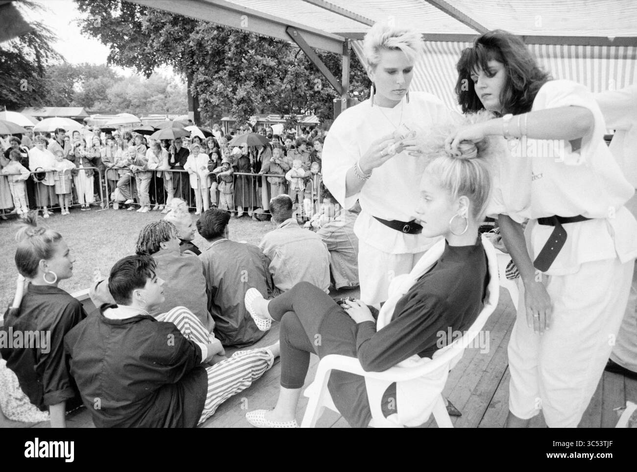 Sfilata di moda e parrucchiere a IJmuiden, Fashion, IJmuiden, Paesi Bassi, 01-07-1988 Whizgle News, Dutch Desk, Paesi Bassi, 1950-2000 in un vivace ambiente all'aperto, gli acconciatori sono visti lavorare sulle acconciature dei modelli come un pubblico accattivante guarda. Gli stilisti mettono in mostra le loro abilità, con uno che prepara sapientemente i capelli di un modello seduto, mentre altri attendono pazientemente il loro turno. L'atmosfera è vivace, piena di emozioni e creatività. Foto Stock